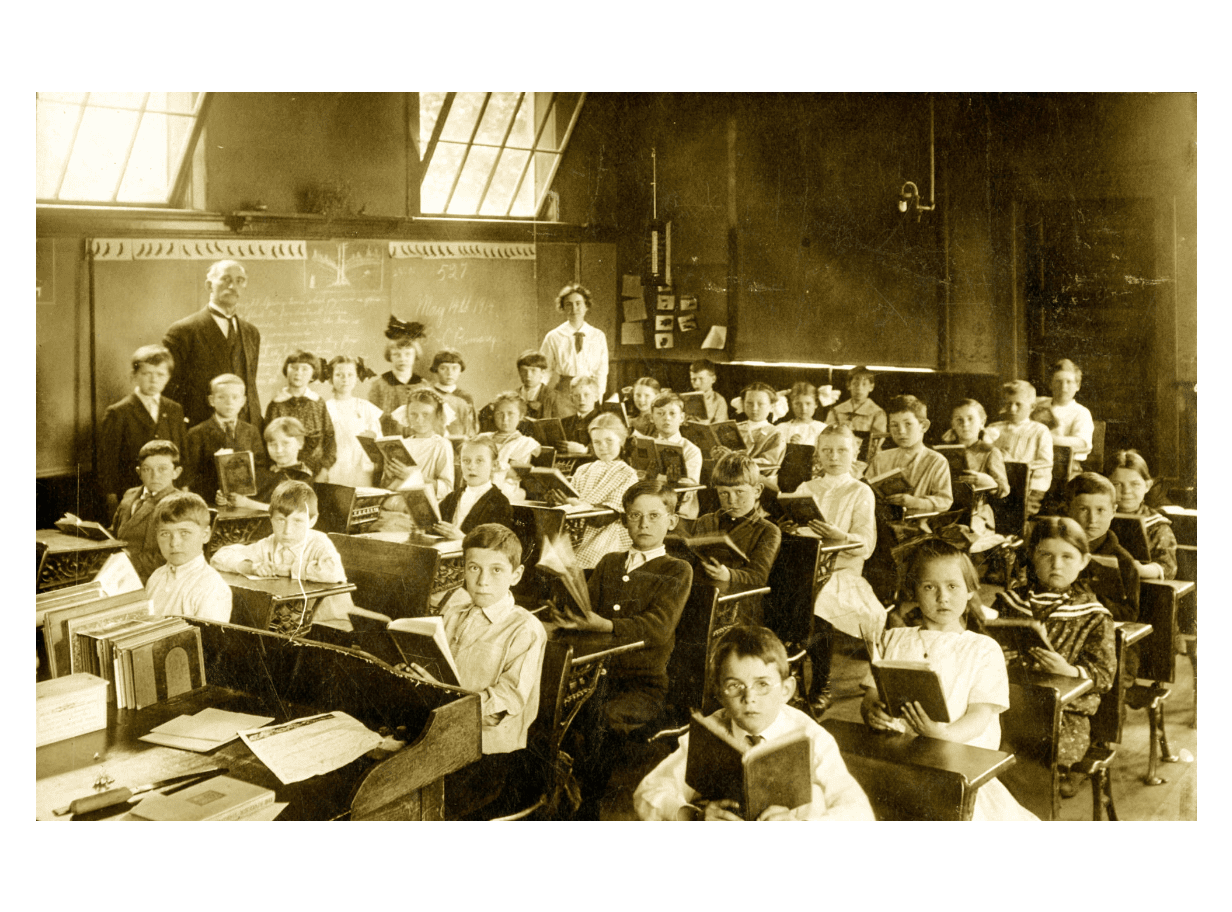 An albumen print of an unidentified classroom of elementary-age children sitting at their desks with their textbooks in hand while their teachers look on