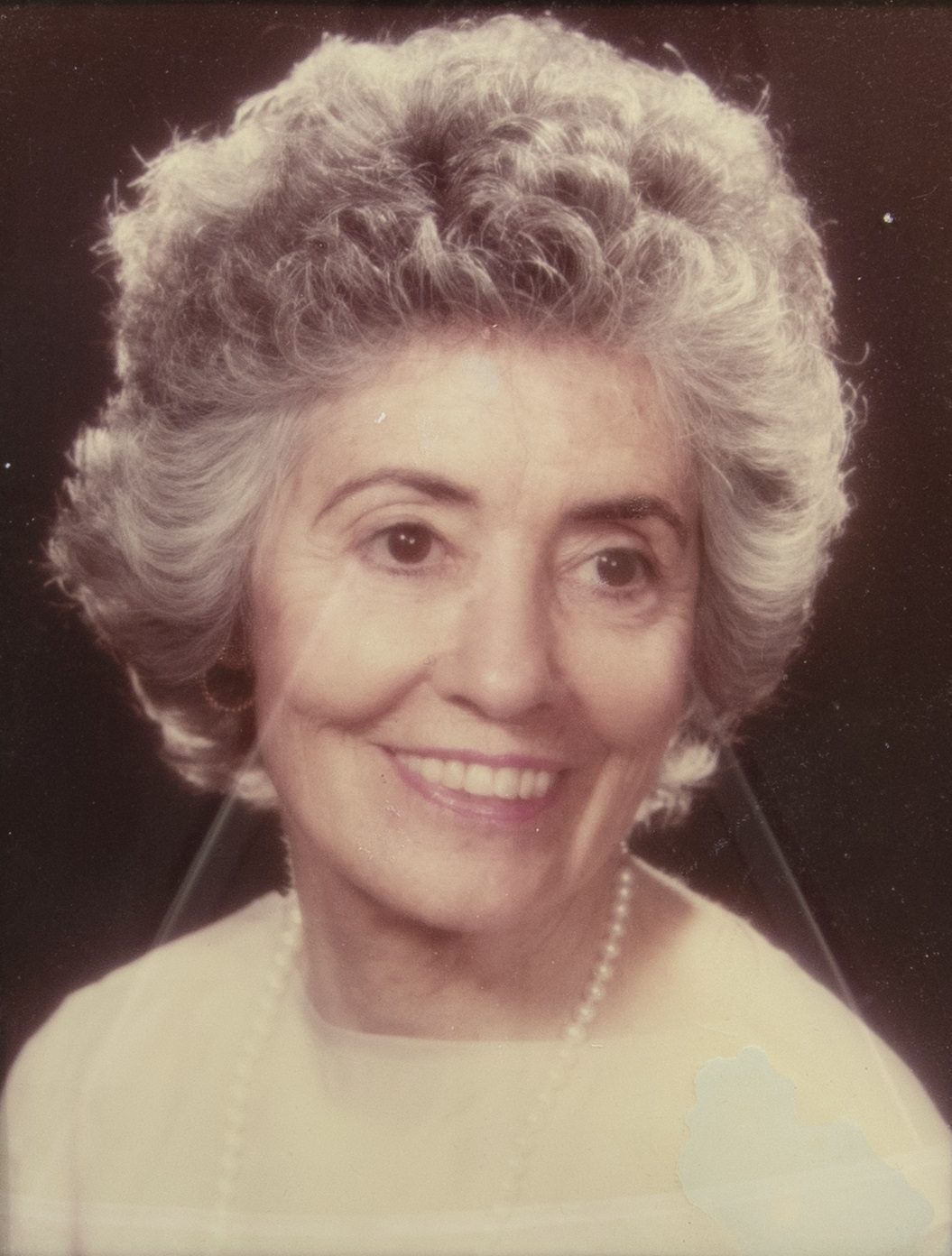 Color photographic portrait of a gray-haired woman wearing a white shirt, a string of pearls, and pearl earrings who smiles at the camera