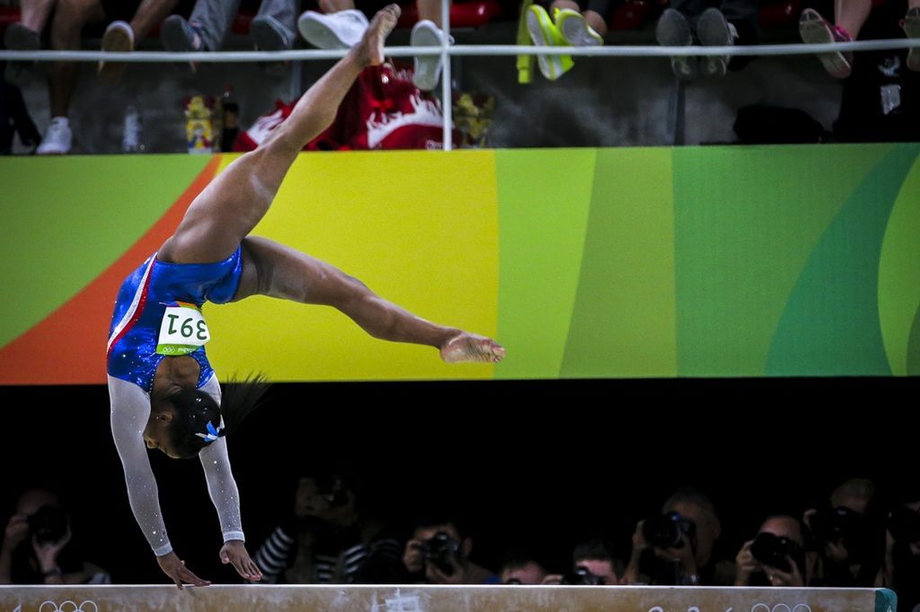 Simone Biles bends her legs backward over her head while balancing on both hands on a balance beam