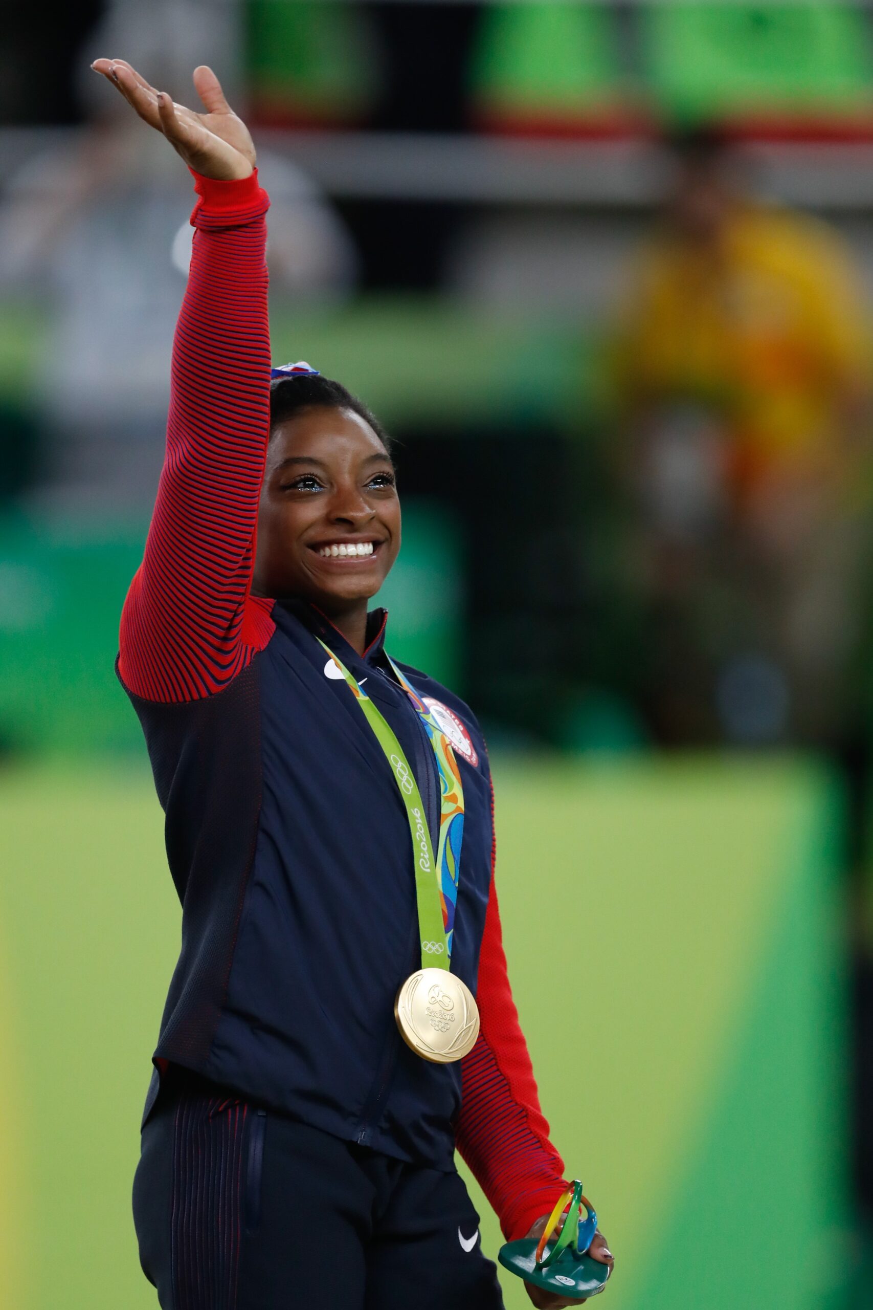 Olympian Simone Biles wears a gold medal and waves with her right hand while smiling