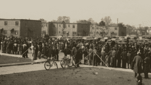 Black and White photo of a large crowd of people standing on the sidewalk and in the parking lot on the