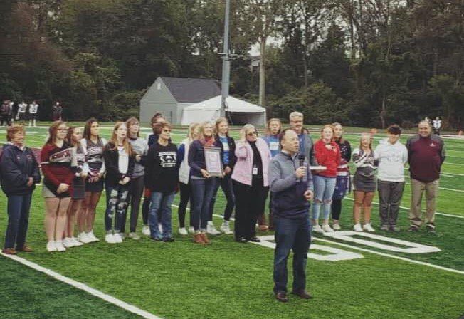 An outside image of a High School football field with students and families standing behind a man with a microphone