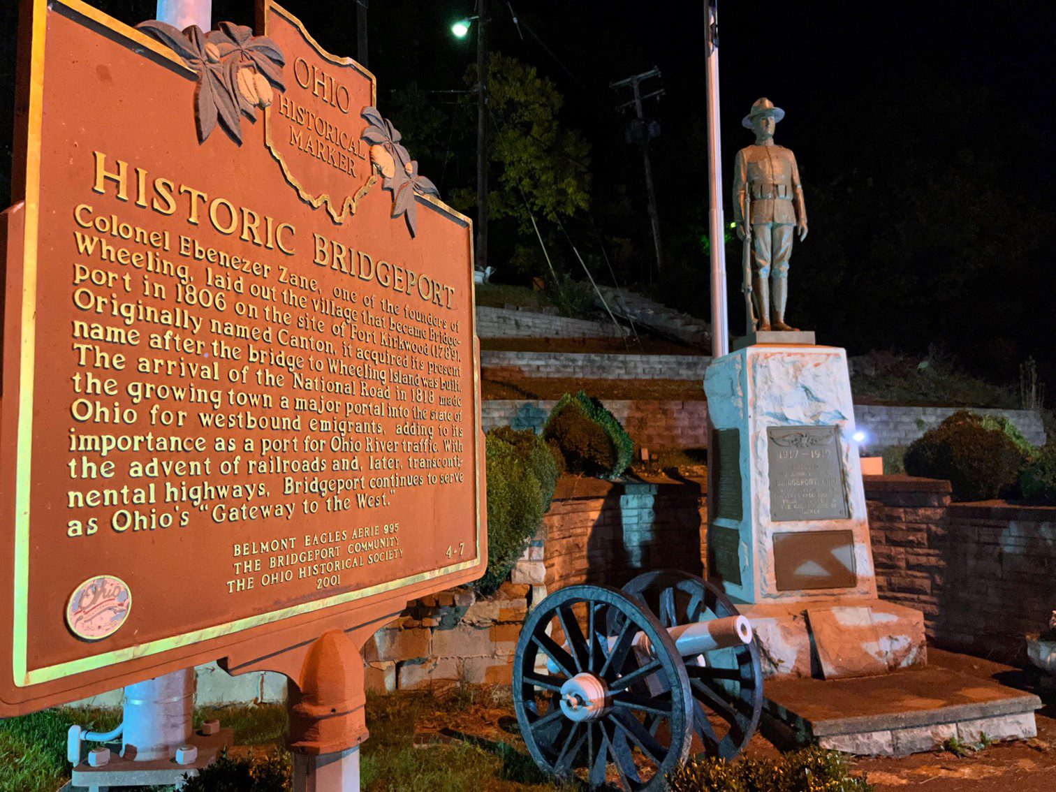 An outdoor image at night with a close-up of a Ohio Historical Marker for Historic Bridgeport. It explains the significance of the area and a statue of a WWI solder.