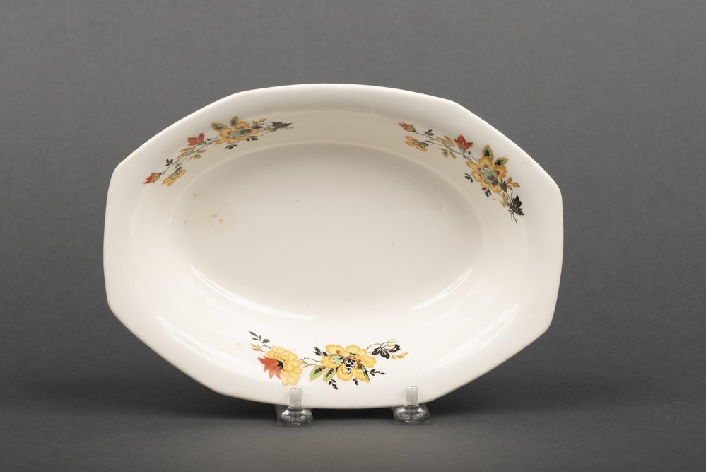 An oval earthenware vegetable dish. The ends of the dish project as flat handles. The plate is decorated with three floral designs in black, yellow, orange and green. The plate was made by homer Laughlin in 1930. 