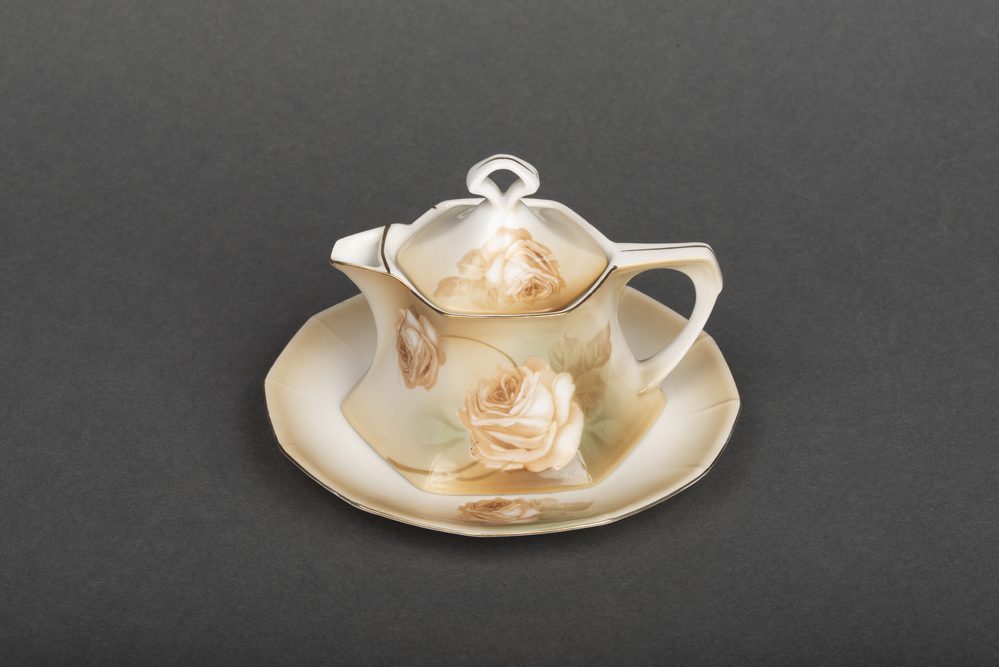 A white, tan and brown porcelain pitcher with gold accents. The pitcher and lid have hexagonal bases. Both the pitcher and its matching saucer are decorated with roses. The porcelain was made in Germany. 