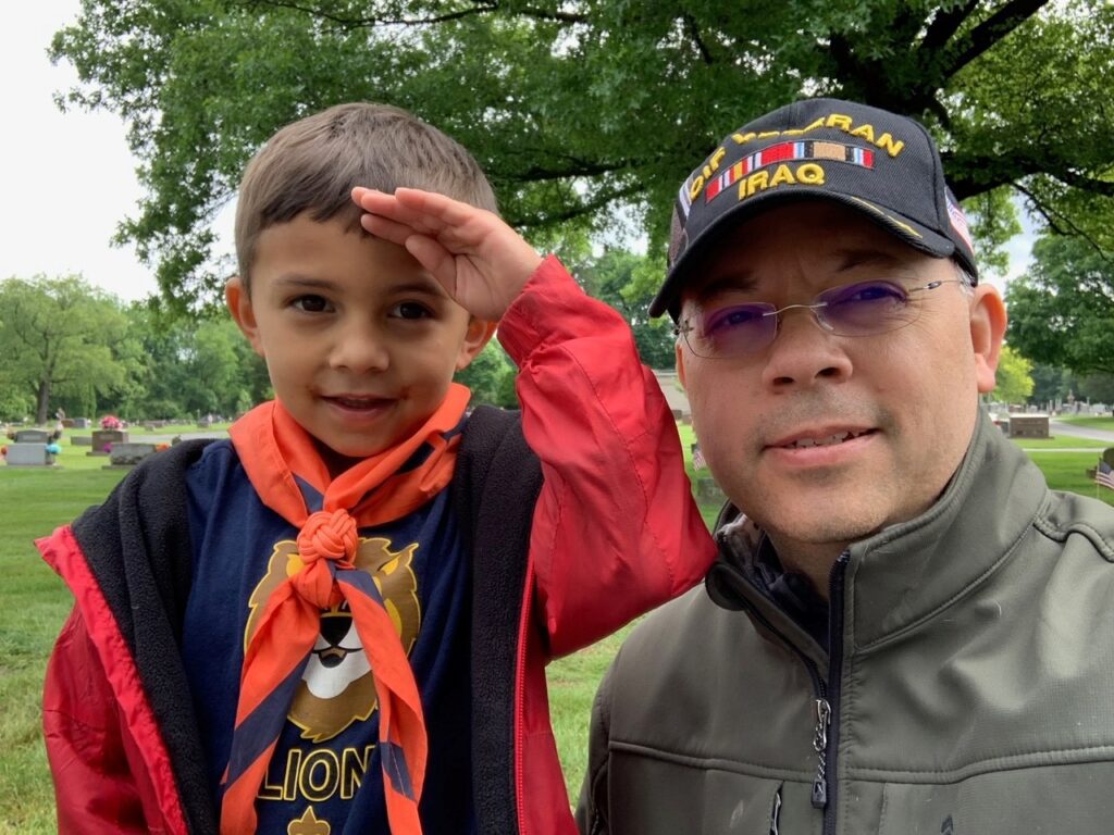 A man is kneeling next to a boy in a Scout uniform who is saluting the photographer.