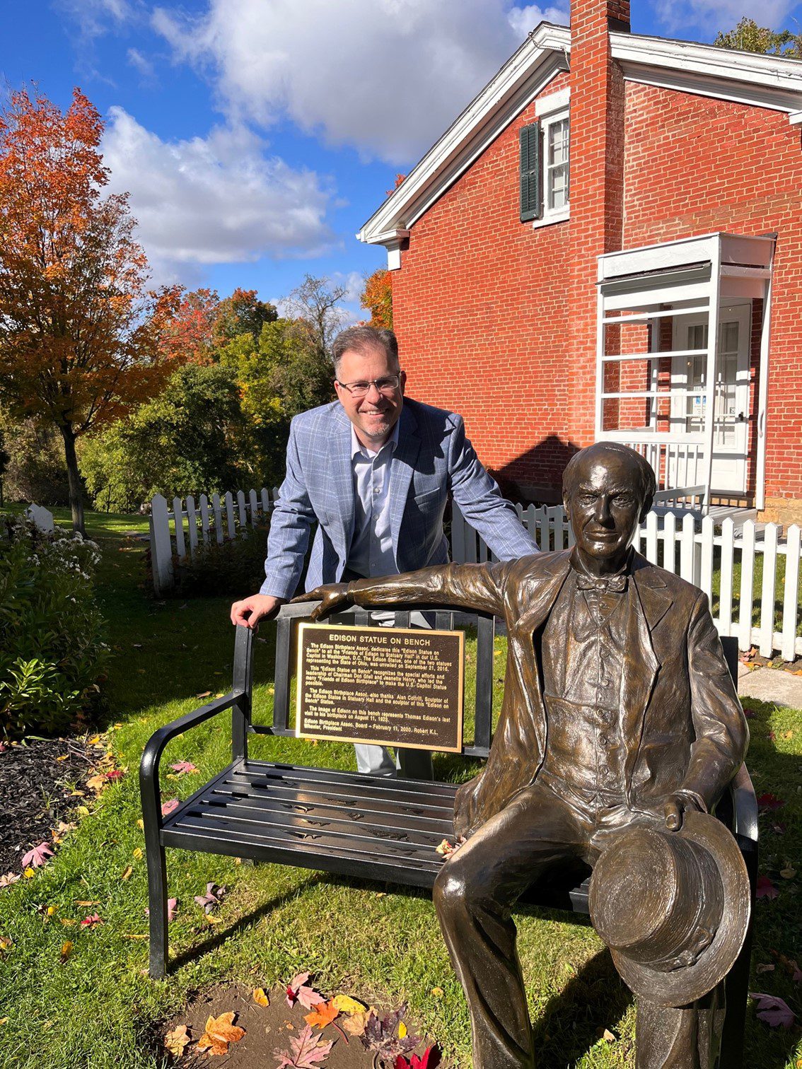 An outdoor photo taken during the day with a white male in a light grey suit who is leaning on a black bench and smiling. He is joined by a dark bronze statue of Thomas Edison who is sitting on the bench. Behind them is a two-story brick building.
