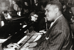 The featured musician, an African American blind male, is playing the piano and his eyes are closed. There are many white audience members in the background.