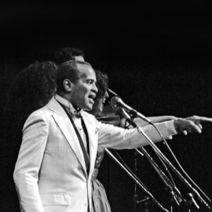 The featured musician, an African American male, is performing on stage with two white women. They are all pointing out to the audience as they sing into the microphones.