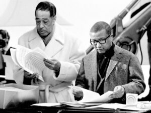Two musicians who are African American males stand next to each other looking at sheet music. They appear to be discussing the arrangement that the featured musician created.