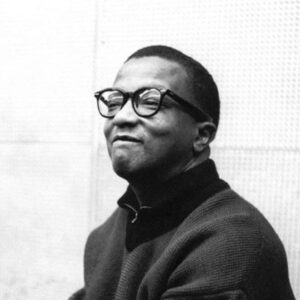 The featured musician, an African American male wearing glasses, looks off to the corner and grins.