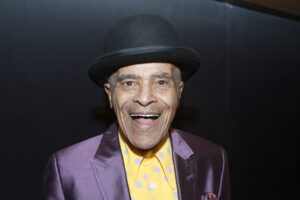 The featured musician, an African American male, looks directly into the camera with a large smile. He is wearing a black top hat, shiny purple suit and a yellow shirt with lavender polka dots..
