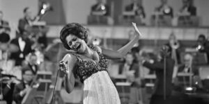 The featured musician, an African American woman, is singing into a microphone with a lot of energy and emotion. She is in front of a large brass band.