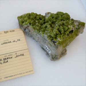 An apple green crystaline mineral alongside a handwritten label with its collection information.