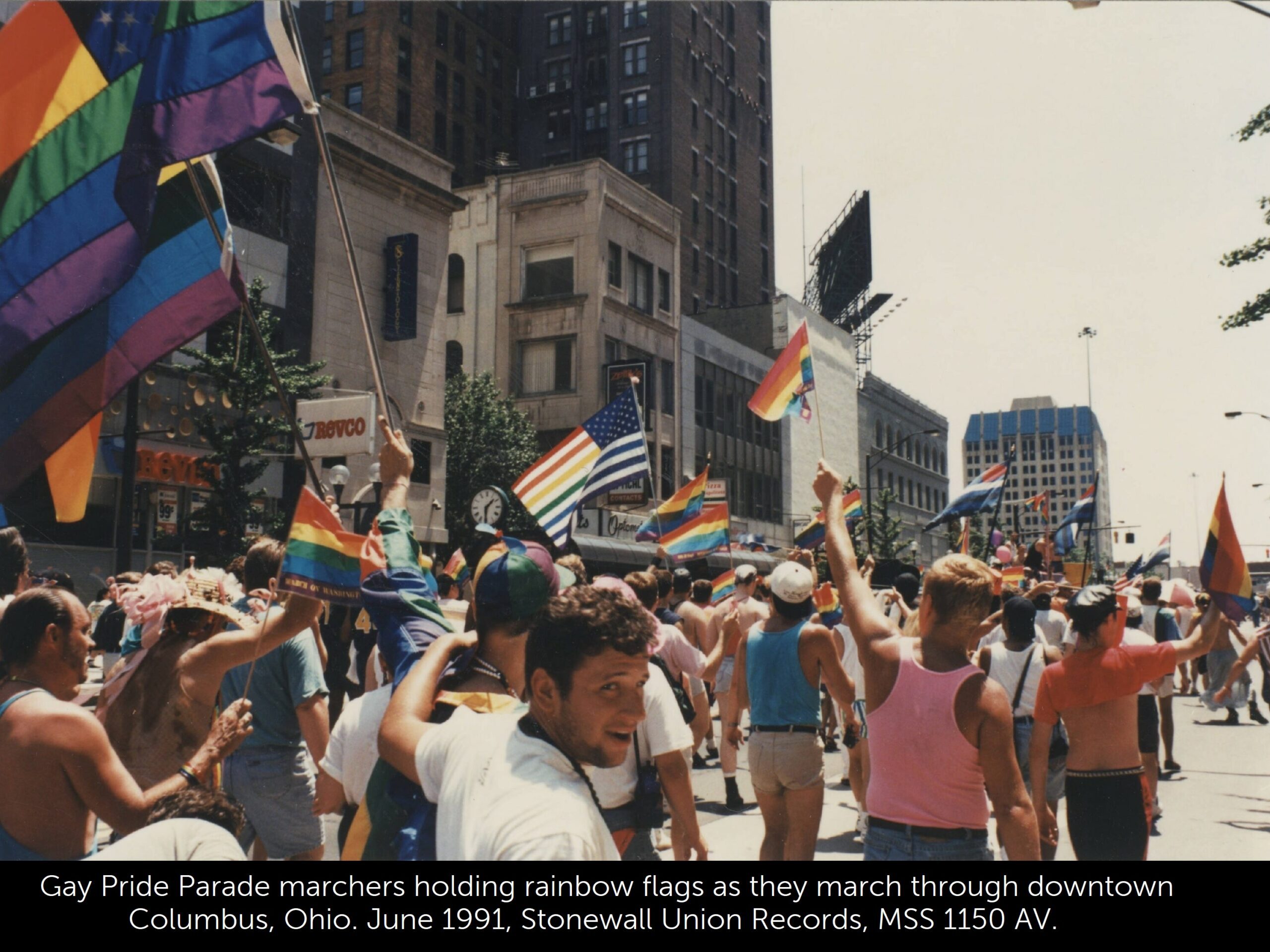 A photo of gay pride marchers holding rainbow flags as they march through downtown Columbus