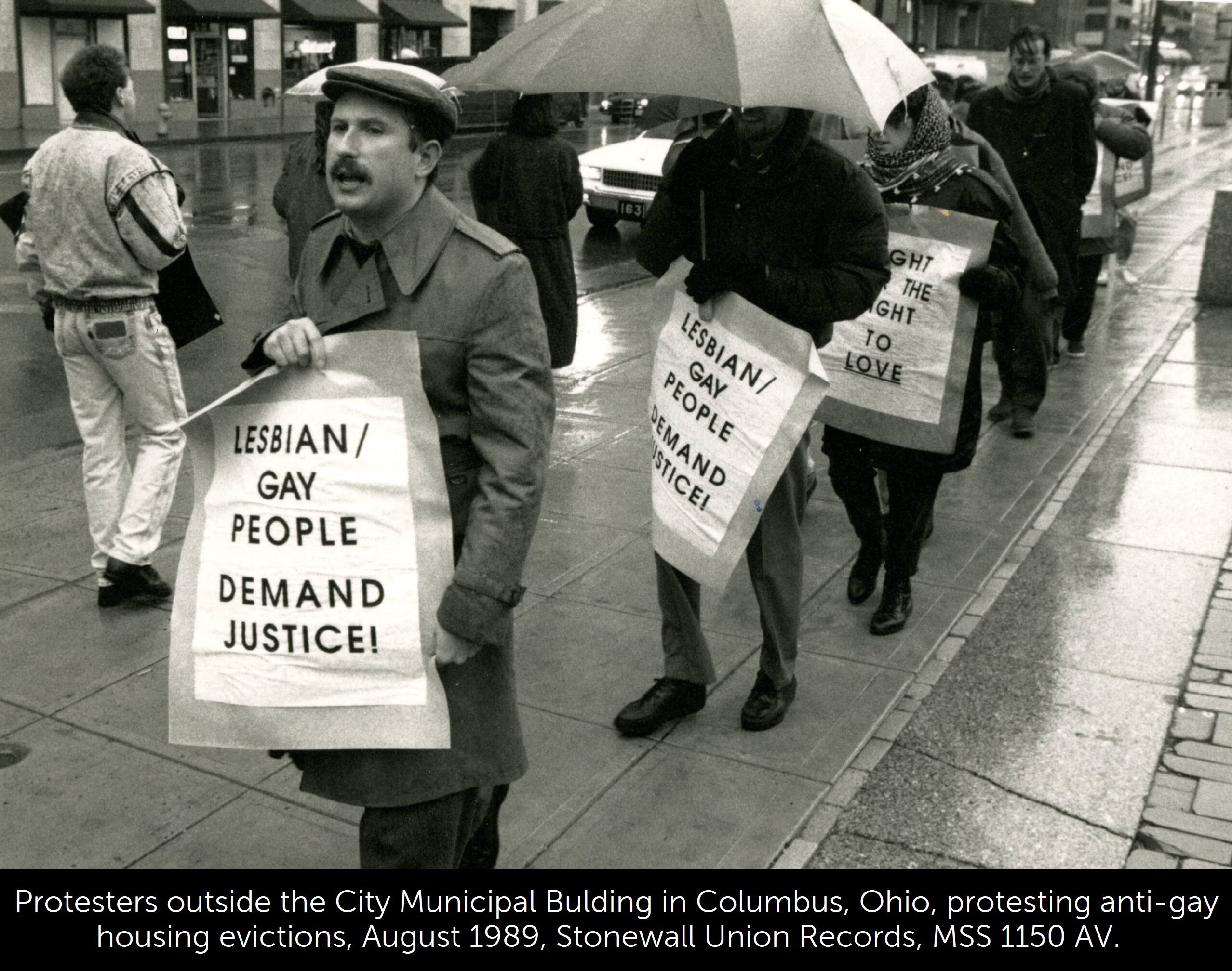 A black and white photo of protesters outside the City Municipal Building in Columbus, holding signs that state, "Lesbian/gay people demand justice!"