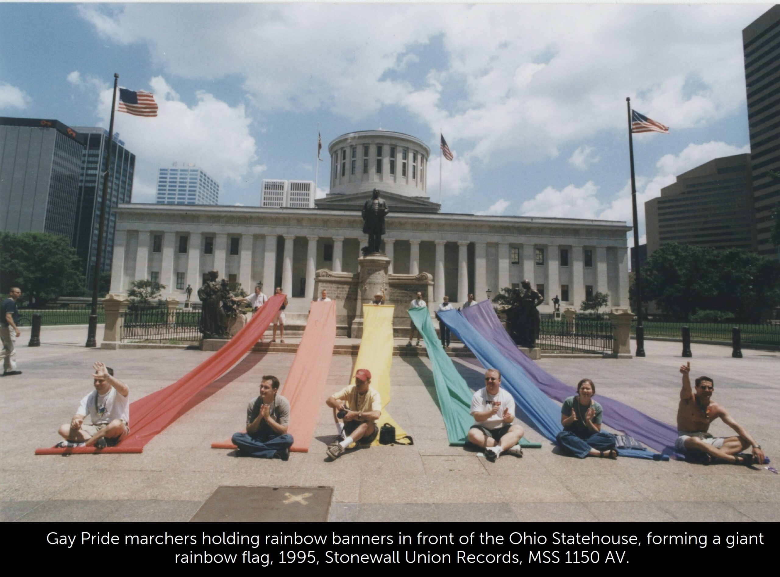 A photo of Gay Pride marchers holding rainbow banners in front of the Ohio Statehouse, forming a giant rainbow flag.