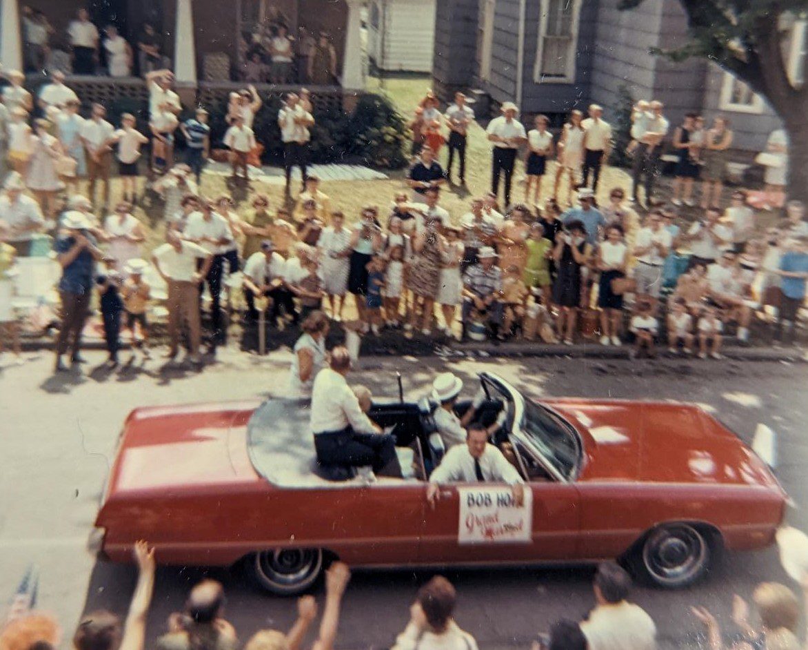 A red convertible driving past crowds of people. A sign on the car reads "Bob Hope Grand Marshal"