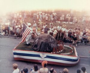 A parade float made in the image of the moon. The American flag is planted on the float. The float is driving past a crowd of people.