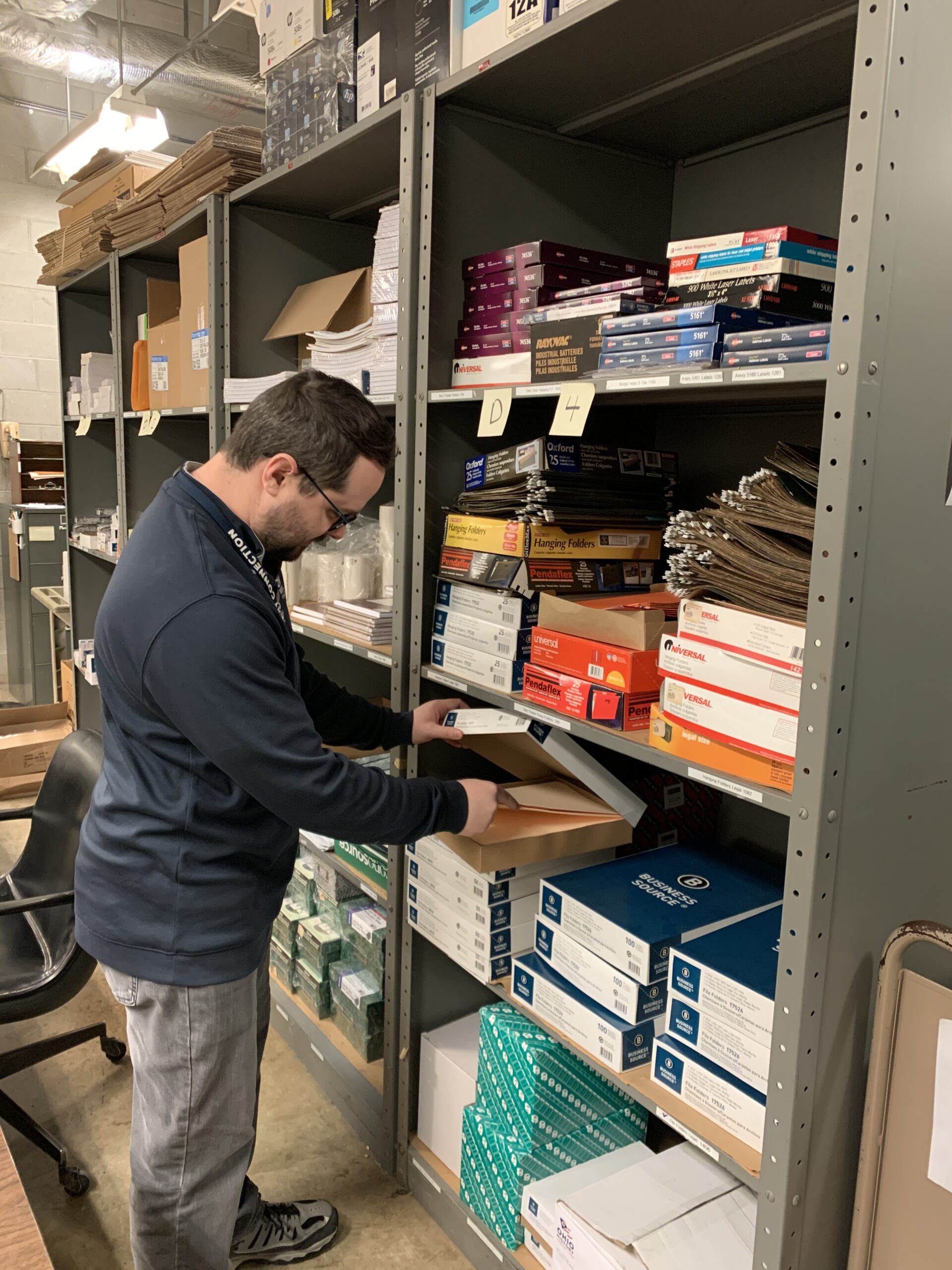 A worker pulling folders out of a box from a shelving unit full of paper products in the distribution center. 