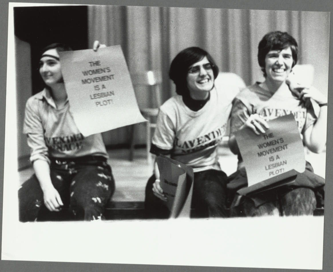 A black and white photo of three lesbian feminist activists son the stage of the Second Congress to Unite Women on May, 1970. They are wearing shirts that read "Lavender Menace" and signs that read "The women's movement is a lesbian plot!"