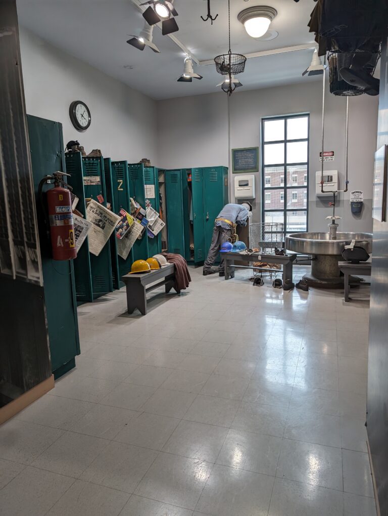 A room with lockers lining the left side. A bench with hard hats on it. A man drinking from a water fountain.