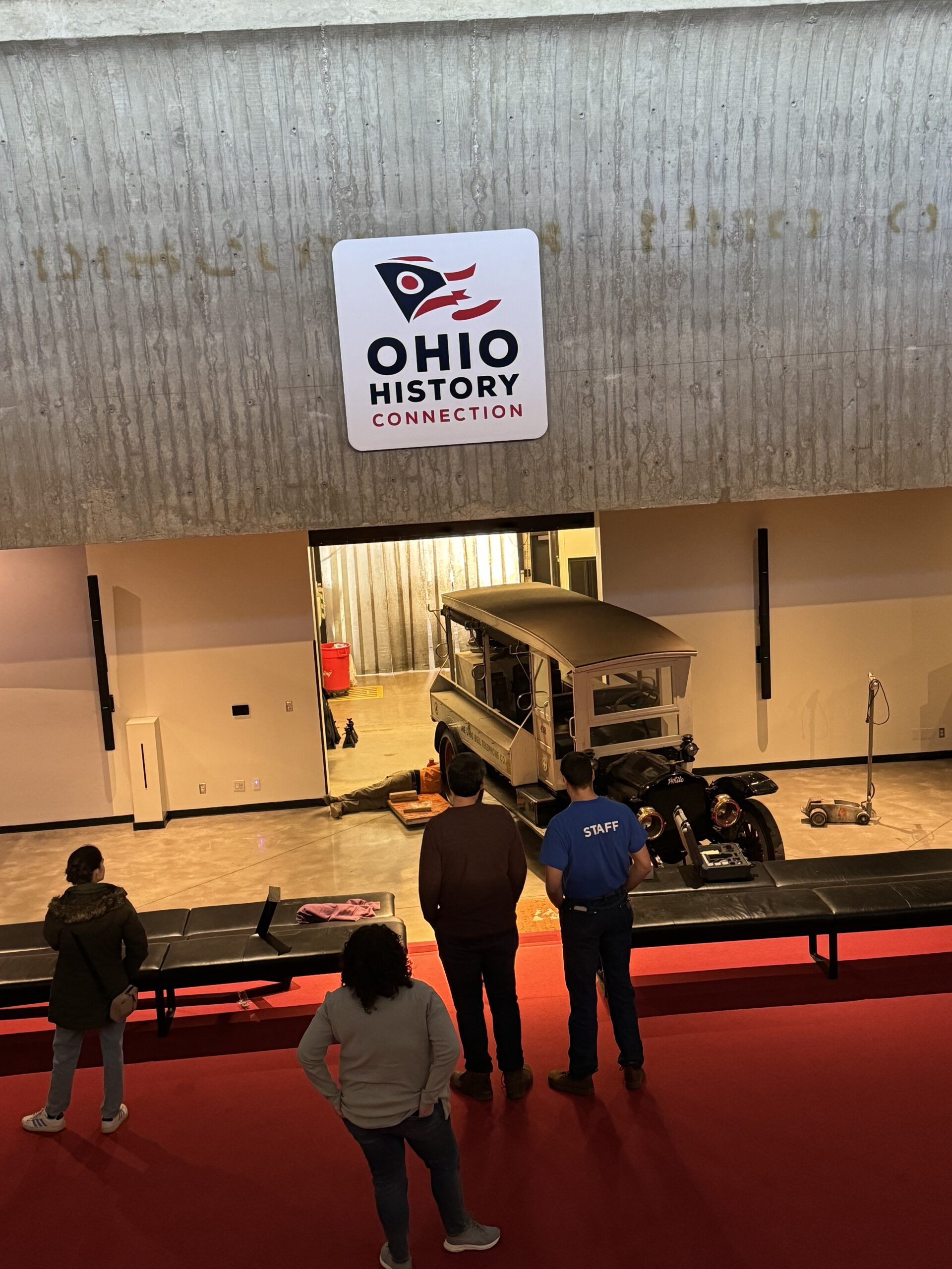 Four people watching an old truck coming in through a loading door to the museum floor. 