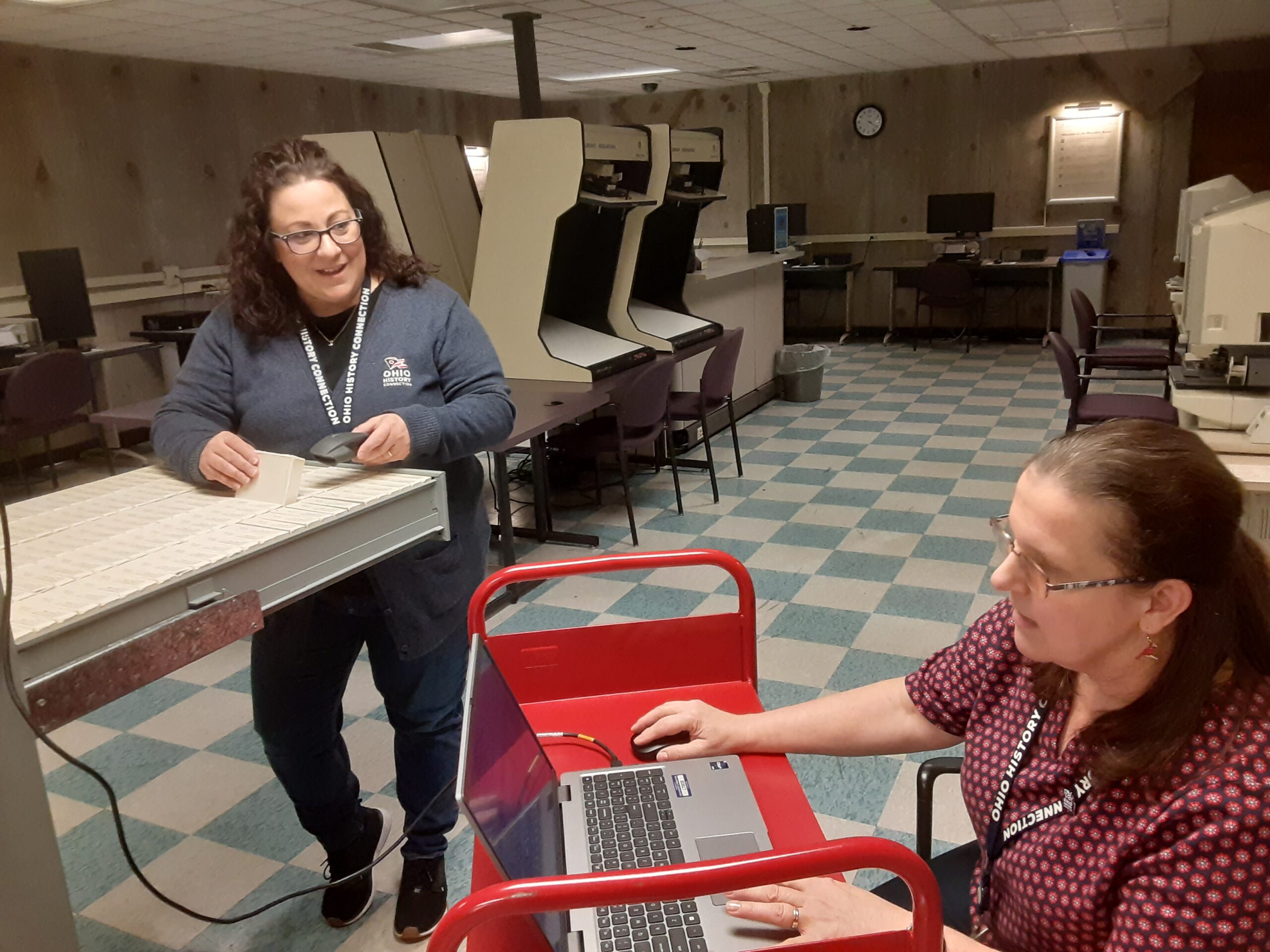 One individual sitting at a computer and a second standing scanning microfilm containers from a storage unit.