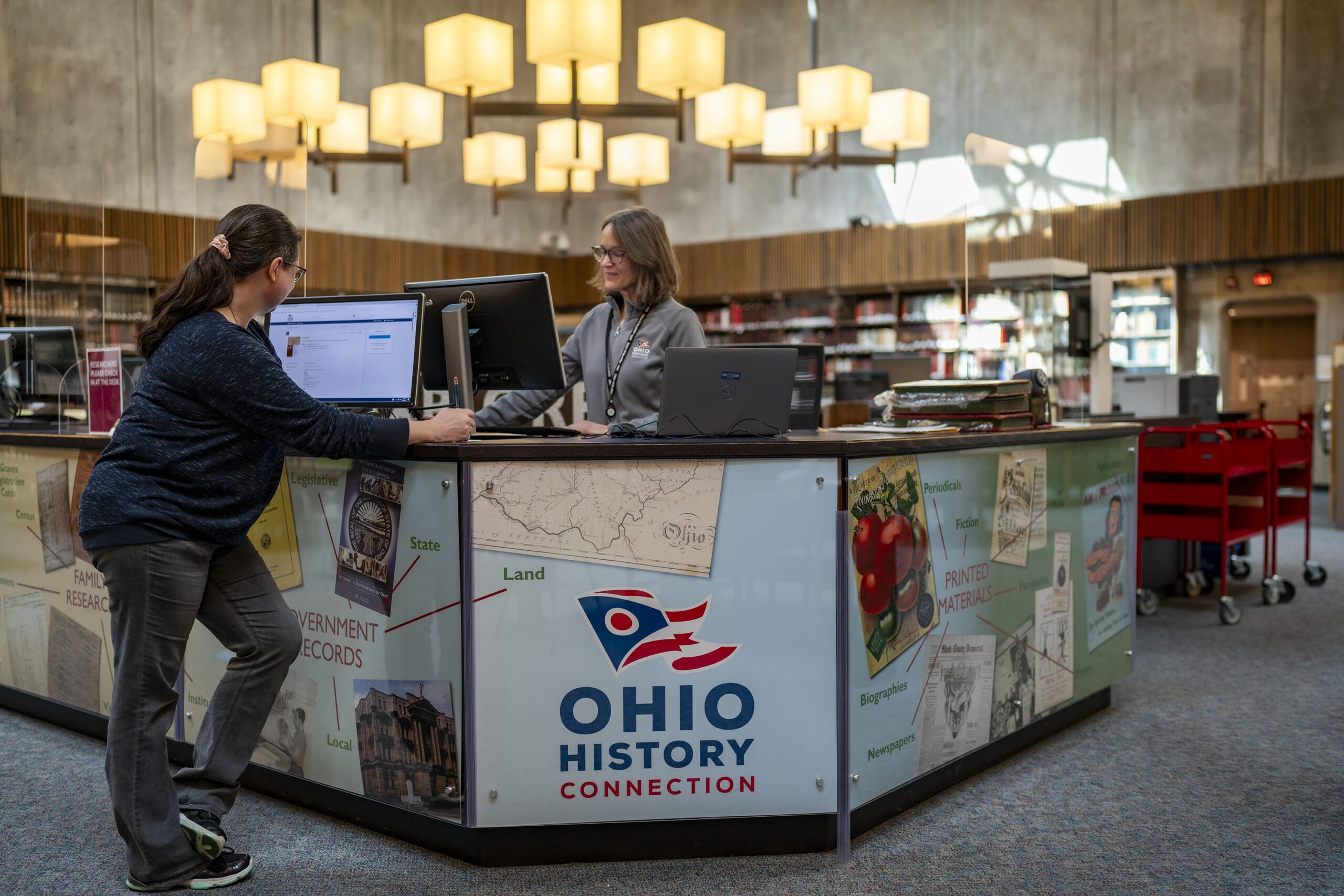 One person standing at a computer behind a welcome desk with another individual in front of her on the other side of the desk.