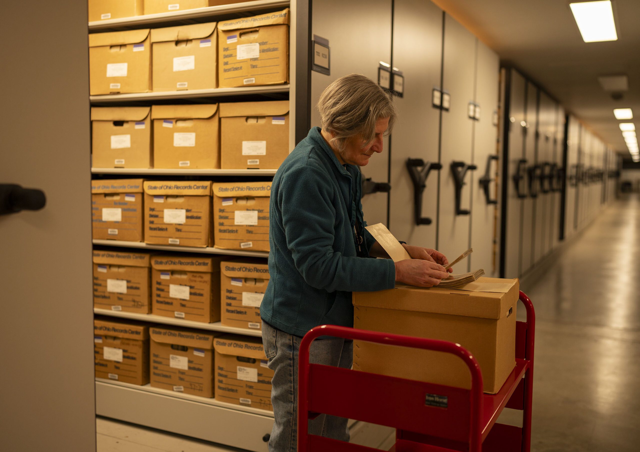 I woman looking through a stack of cards in an aisle between shelves of boxes.