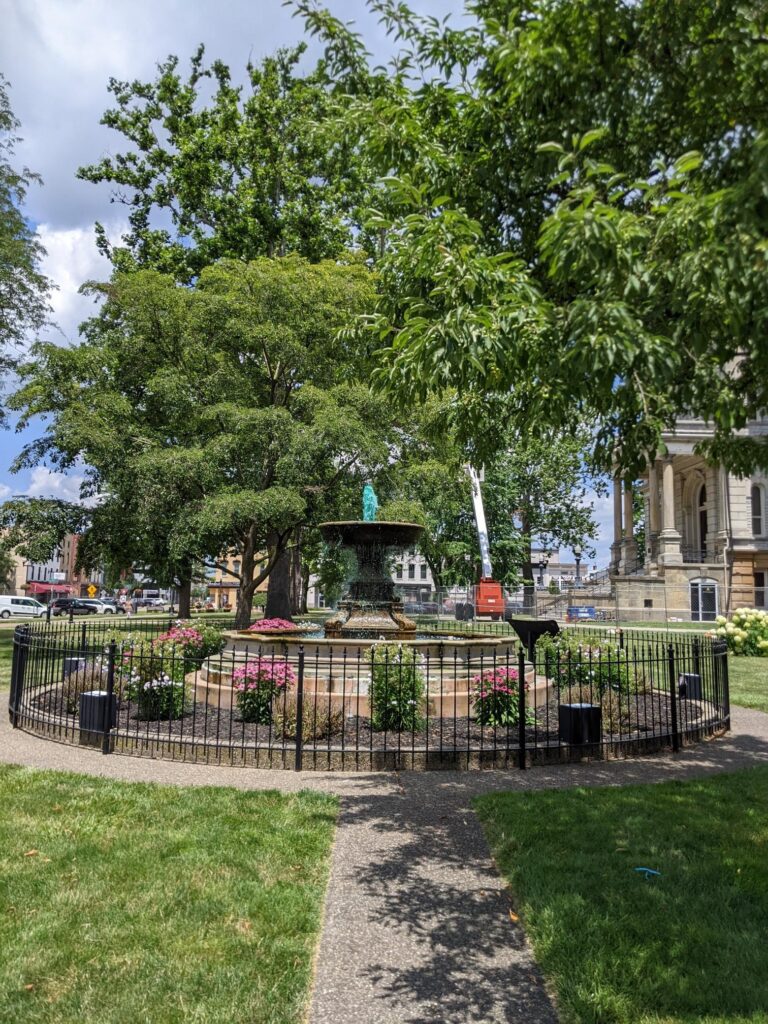 Image of a large water fountain surrounded by garden beds with pink and white flowers planted.