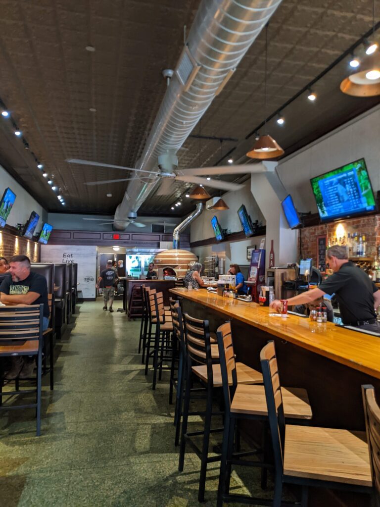 Image of an interior of a restaurant on the right side there is a bar top with chairs and 2 people behind the bar. The left side of the image is tables with seating for customers.