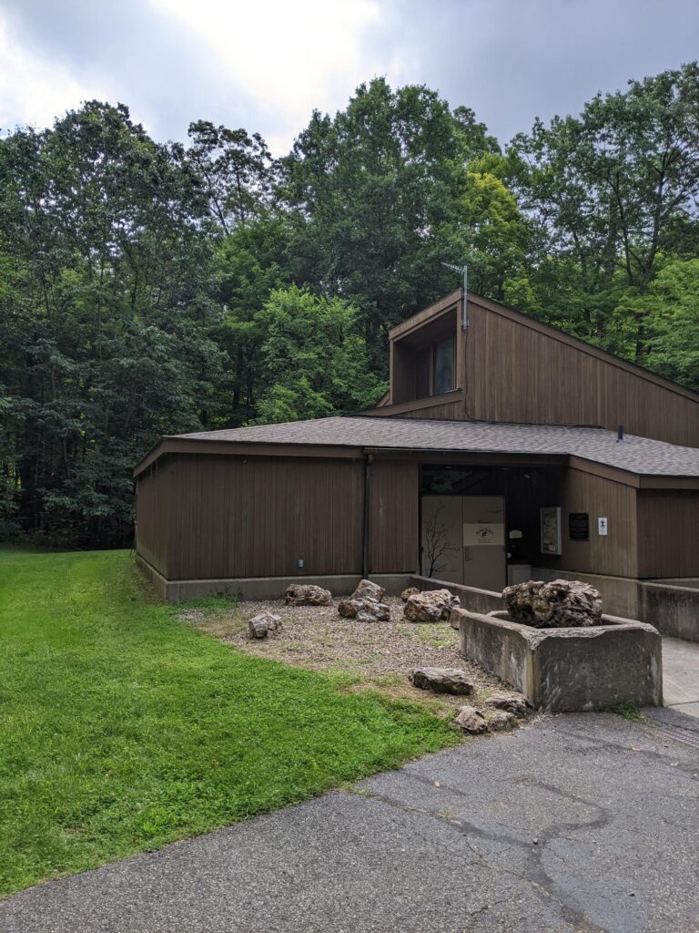 Image of a large brown sided building with large stones out front and trees in the background.