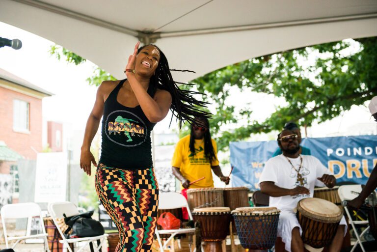 Woman dancing at Poindexter Village Drum Circle, with drummers playing in the background. She is wearing black, gold, green and red patterned pants and a coordinating top.