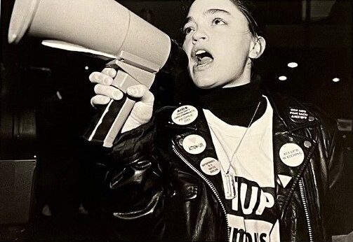 Black and white photograph of activist speaking into a bullhorn in a leather jacked with ACT UP pins