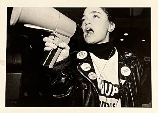 Black and white photograph of activist speaking into a bullhorn in a leather jacked with ACT UP pins