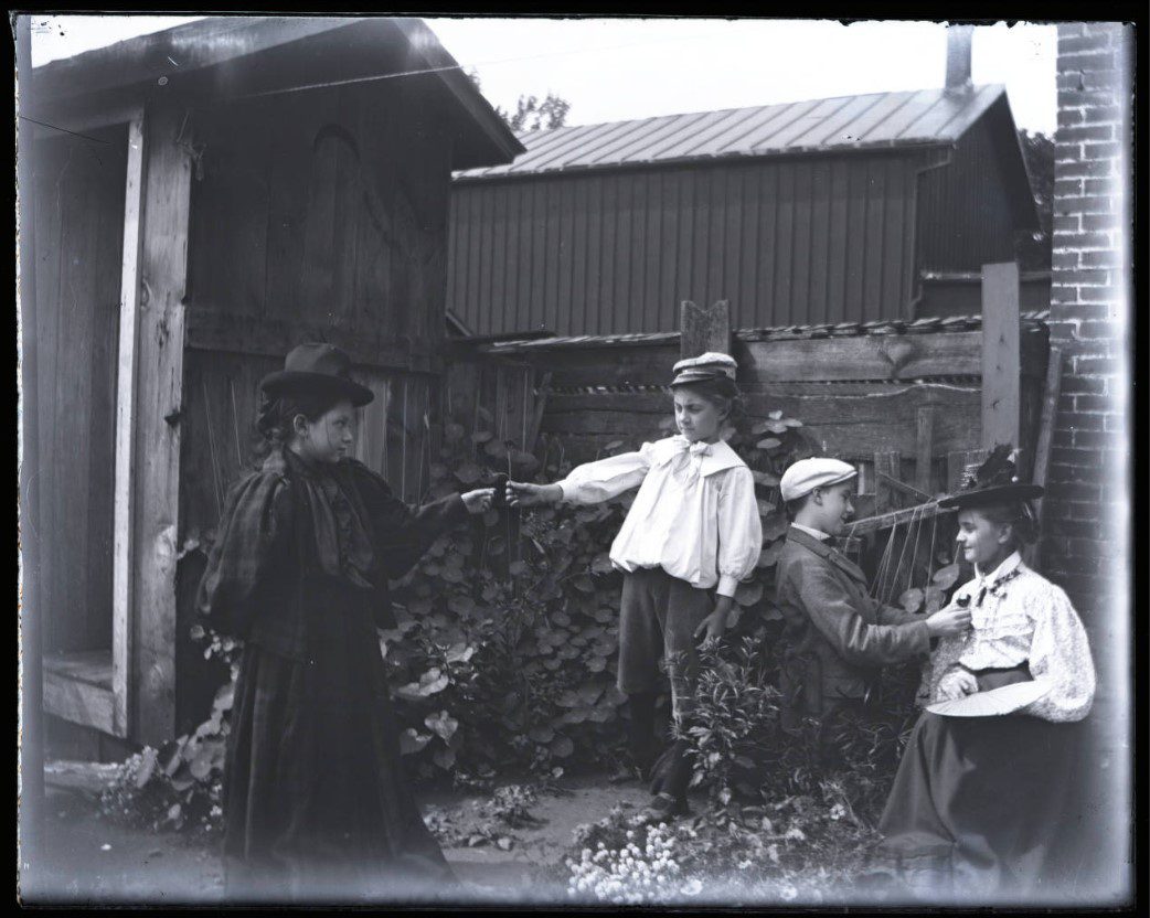 Four children dressed in costumes, playing in front of an outhouse. Two of them are splitting a wishbone.