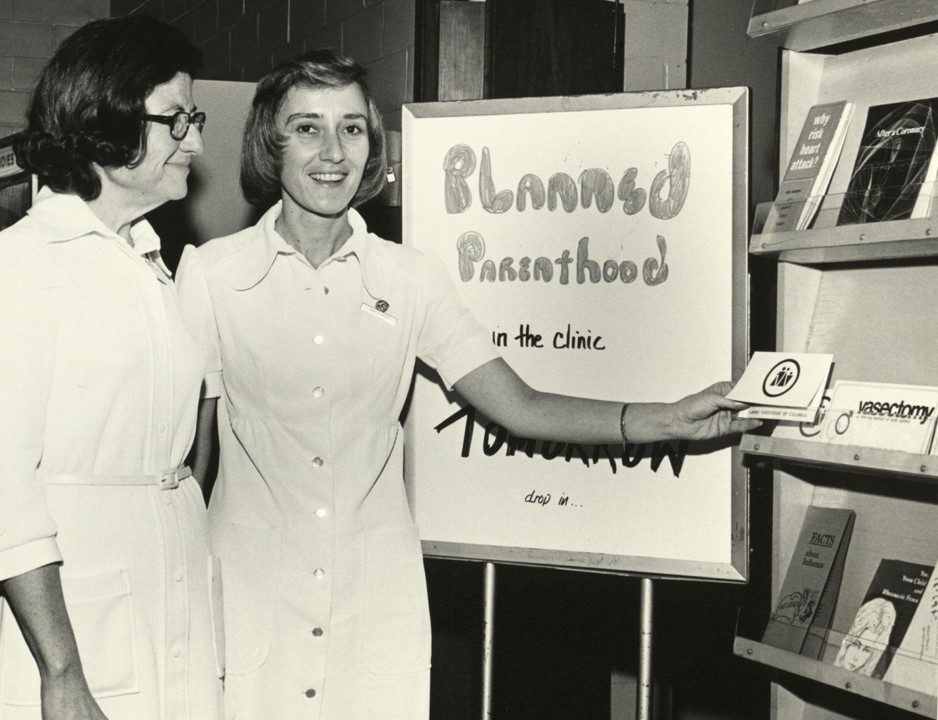 Black and whilte photograph of two women in front of a Planned Parent signboard. One is holding a pamphlet.