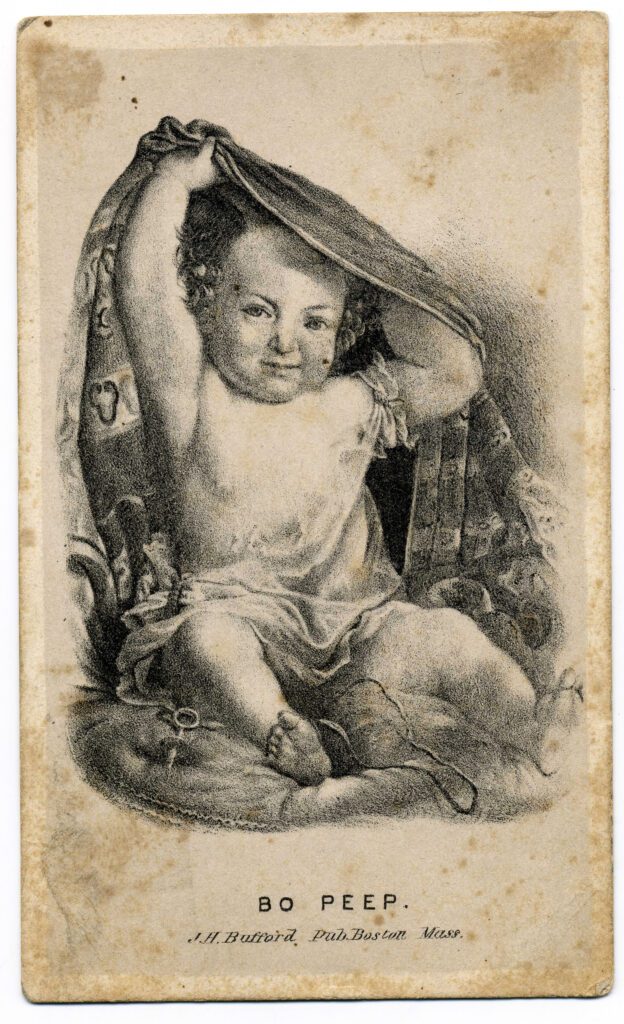 Black and white image of a chubby baby sitting on a pillow pulling a blanket from behind their back and over their head.