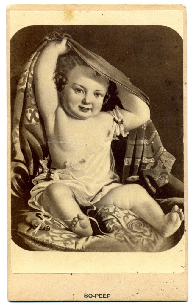Black and white image of a chubby baby sitting on a pillow pulling a blanket from behind their back and over their head.