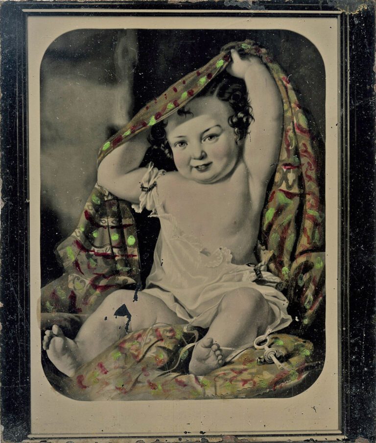 Hand-colored tintype image of a chubby baby sitting on a pillow pulling a blanket from behind their back and over their head.