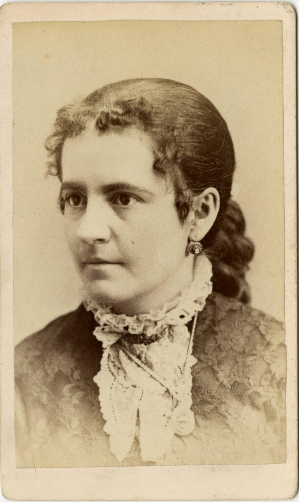 Black and white photographic portrait of a woman with curly hair pulled back from her face wearing a lace dress with a lace collar.