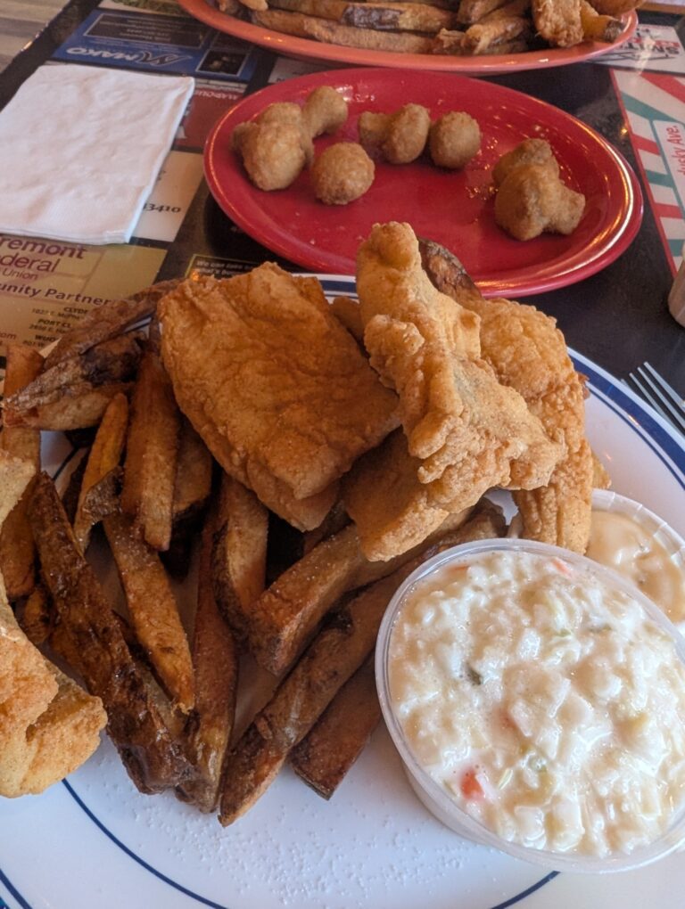 A plate of food containing fries, fried fish, and a bowl of sauce. Fried mushrooms on a plate in the background.