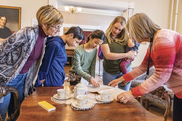 Image of 3 adult women, 2 children, and a baby observing a document on a wooden table in the Harriet Beecher Stowe House. There is also a tea set on the table.