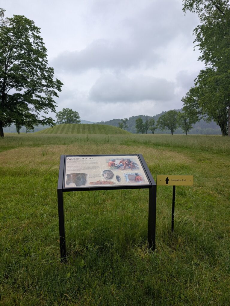 A grassy field showing a portion of hte Seip Earthworks with trees on either side of the photo. There is a sign in the foreground of the photo with information about the site that is not legible.