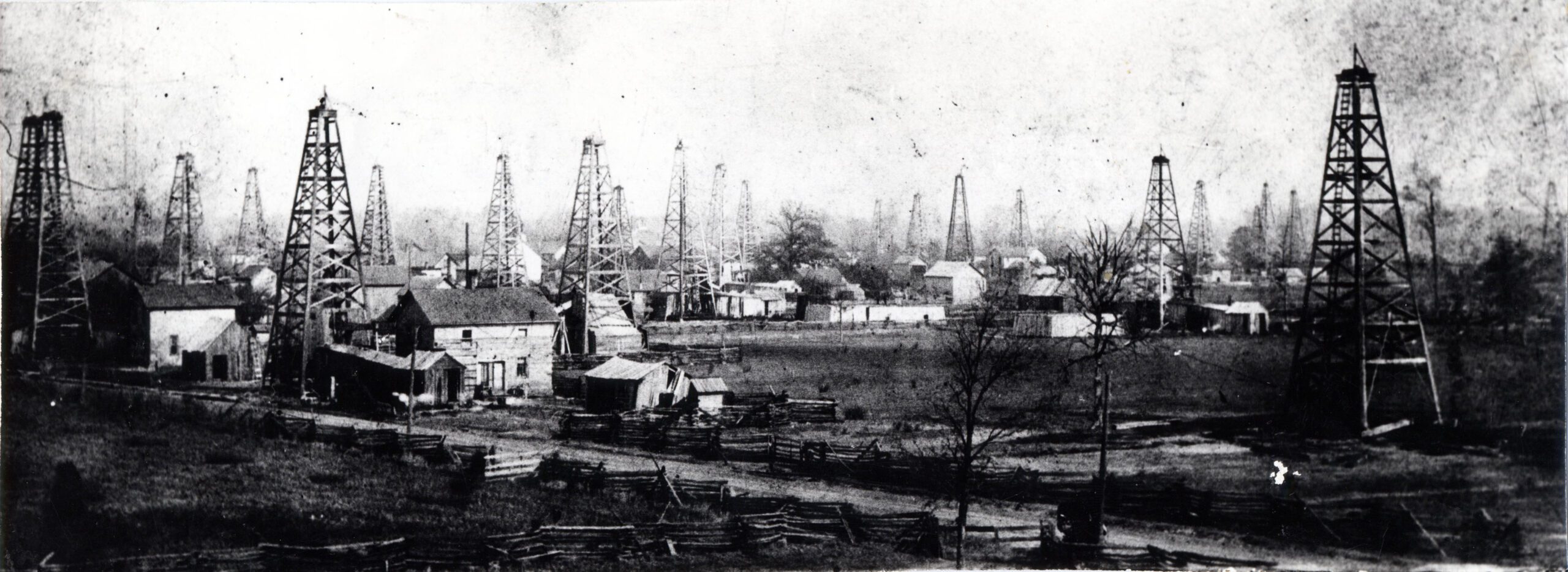 Black and white photograph of two dozen oil derricks. The tall towers protrude from fields, front yards, and even barns in a town.