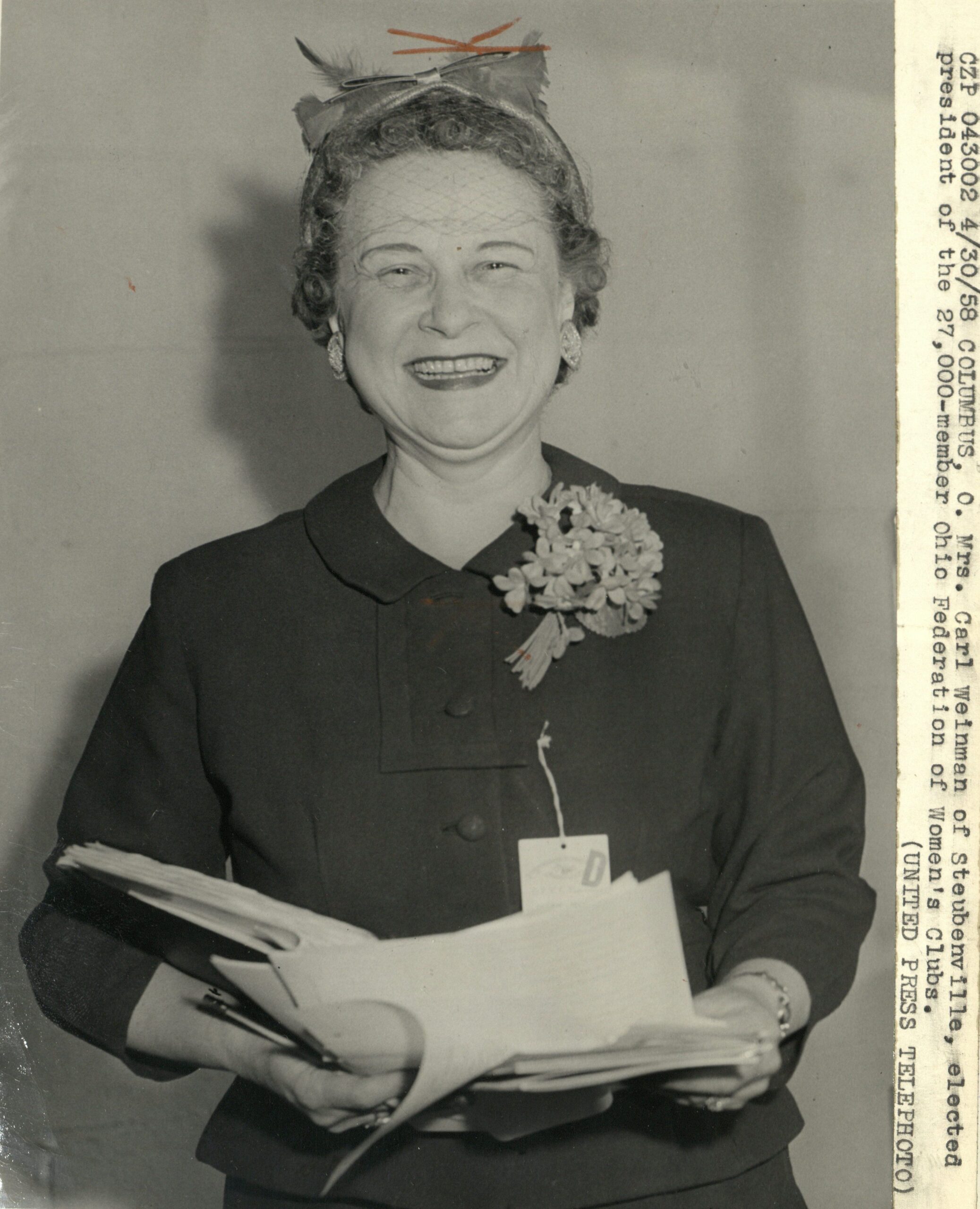Black-and-white photograph of a smiling woman holding papers. A typed label on the right-hand side reads: CZP 043002 4/30/58 COLUMBUS, O. Mrs. Carl Weinman of Steubenville, elected president of the 27,000-member Ohio Federation of Women's Clubs. (UNITED PRESS TELEPHOTO)