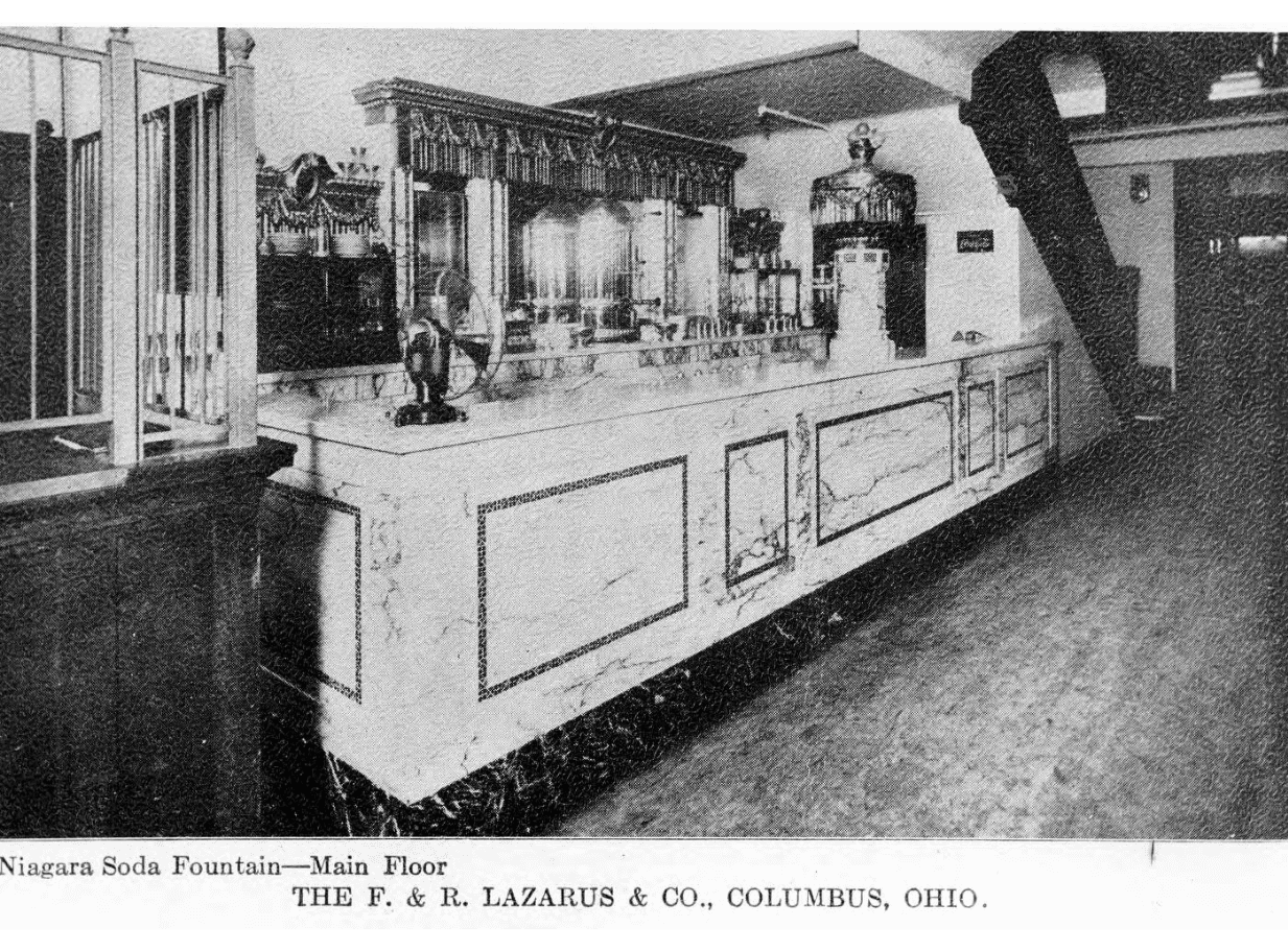 Postcard featuring the Niagara soda fountain at The F. & R. Lazarus Company, Columbus, Ohio, 1909. Image depicts white marble counter set in underneath a staircase. 