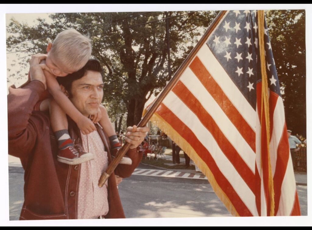 Color photograph of man holding son on his shoulders and an American flag marching in a parade
