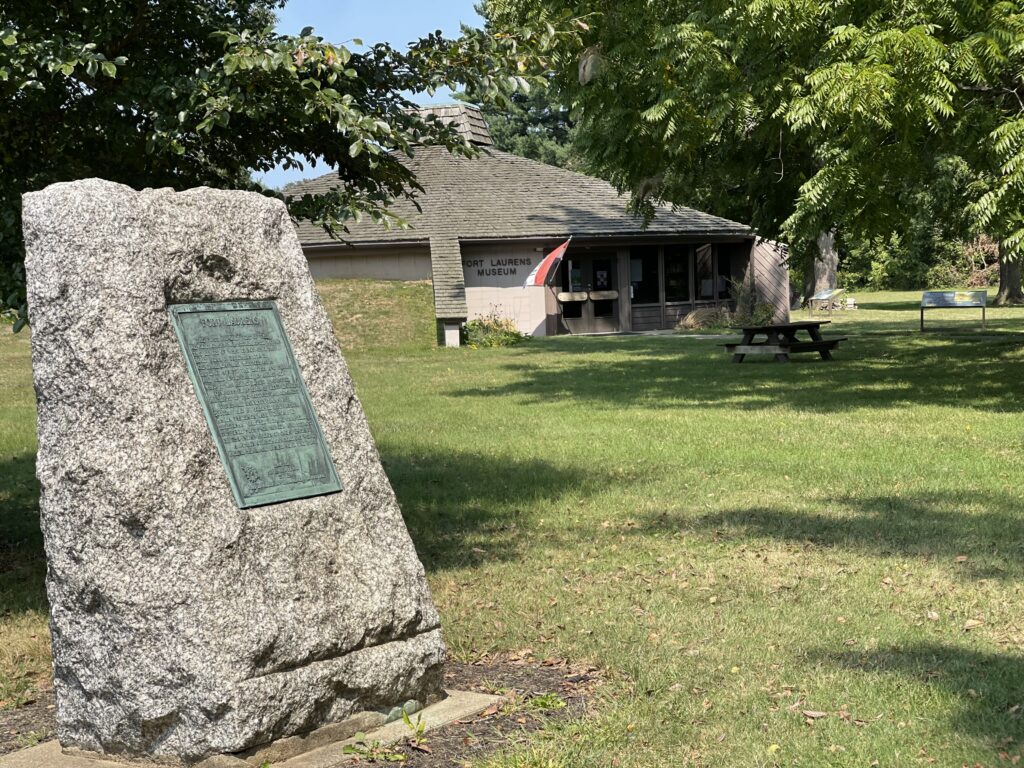 A stone monument stands in the foreground in commemoration to the soldiers at Fort Laurens. The Fort Laurens Museum is in the background.